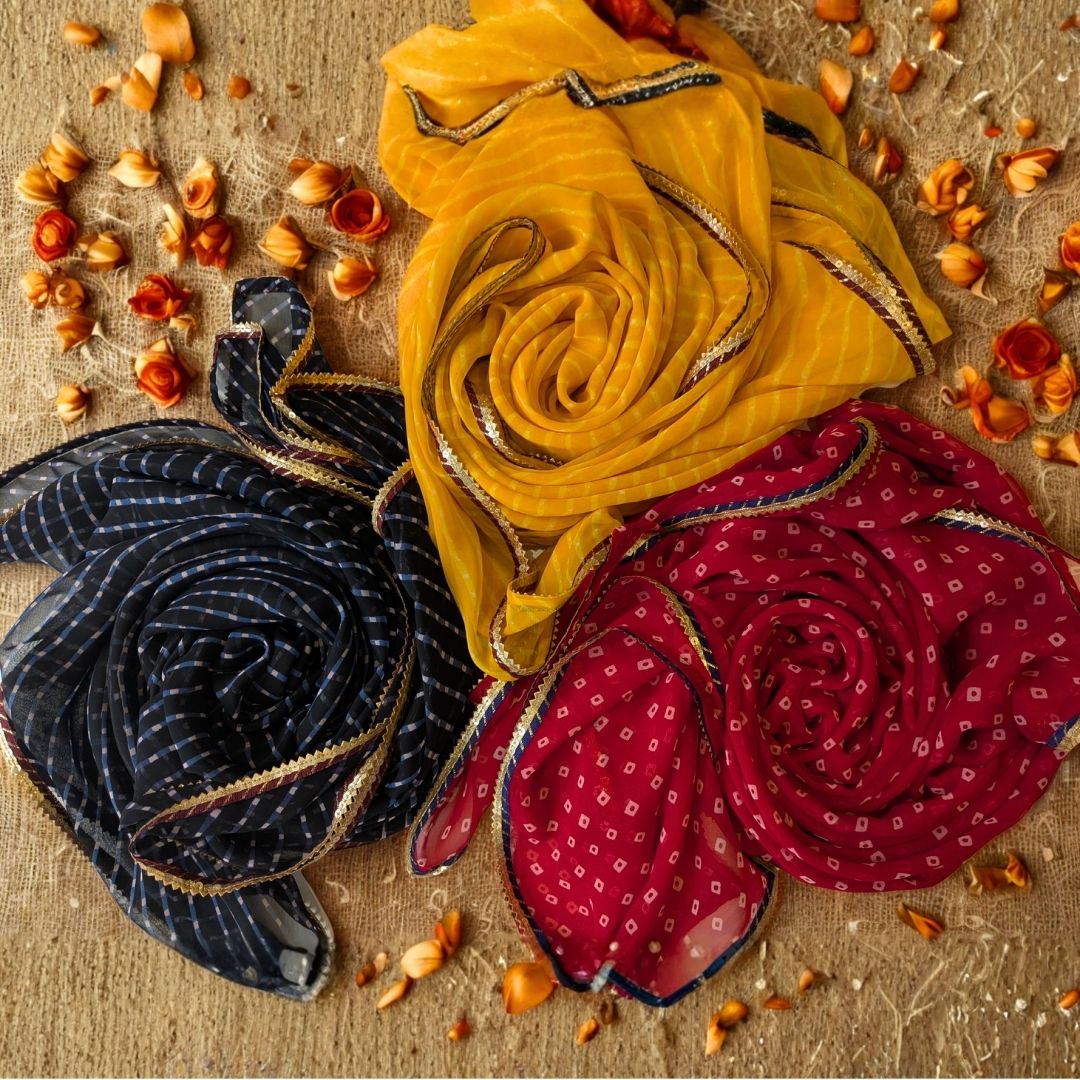 Three rolled-up leheriya dupattas in yellow, black, and red on a textured surface with small flowers.