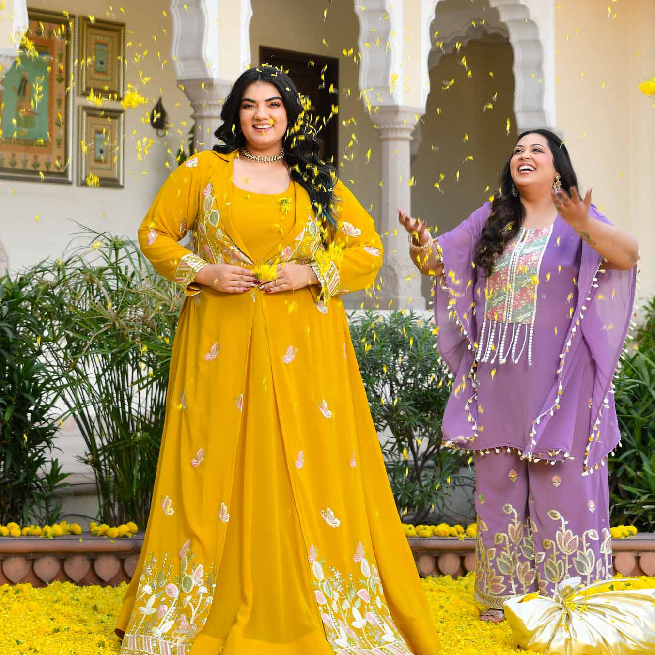 Three women in traditional outfits standing on a path with yellow flowers, in front of an architectural building.